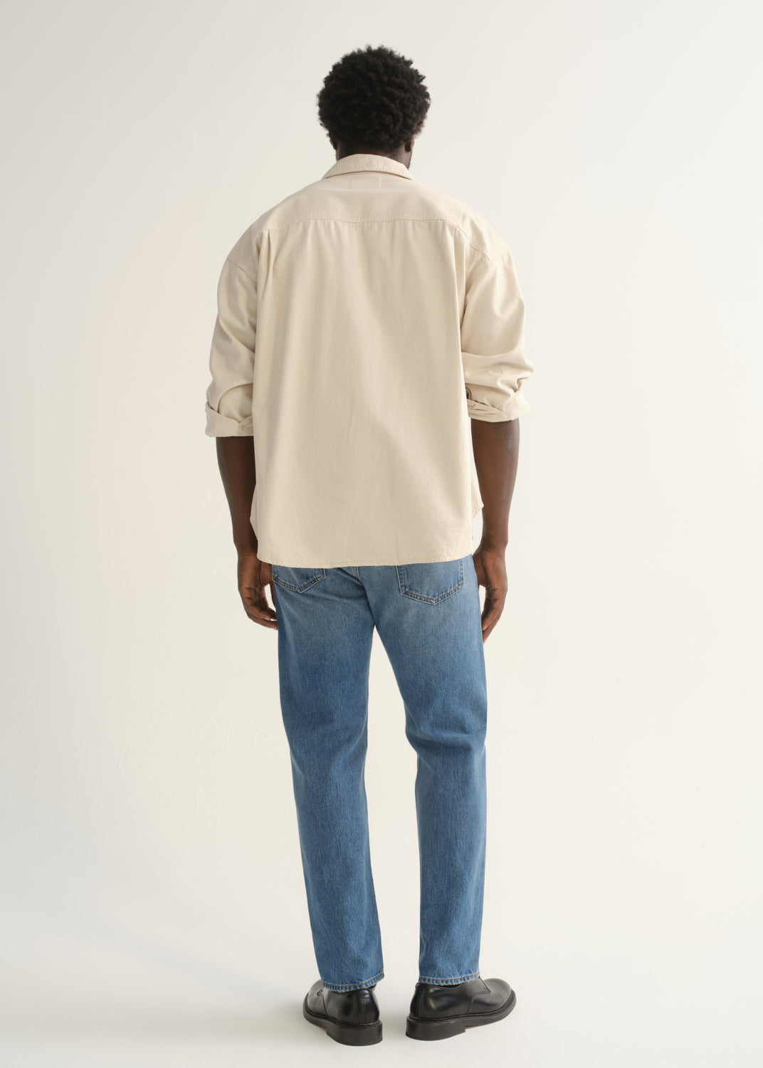 Person wearing light blue straight leg jeans with a cream button-up shirt and black shoes, back full body view on a light grey background.