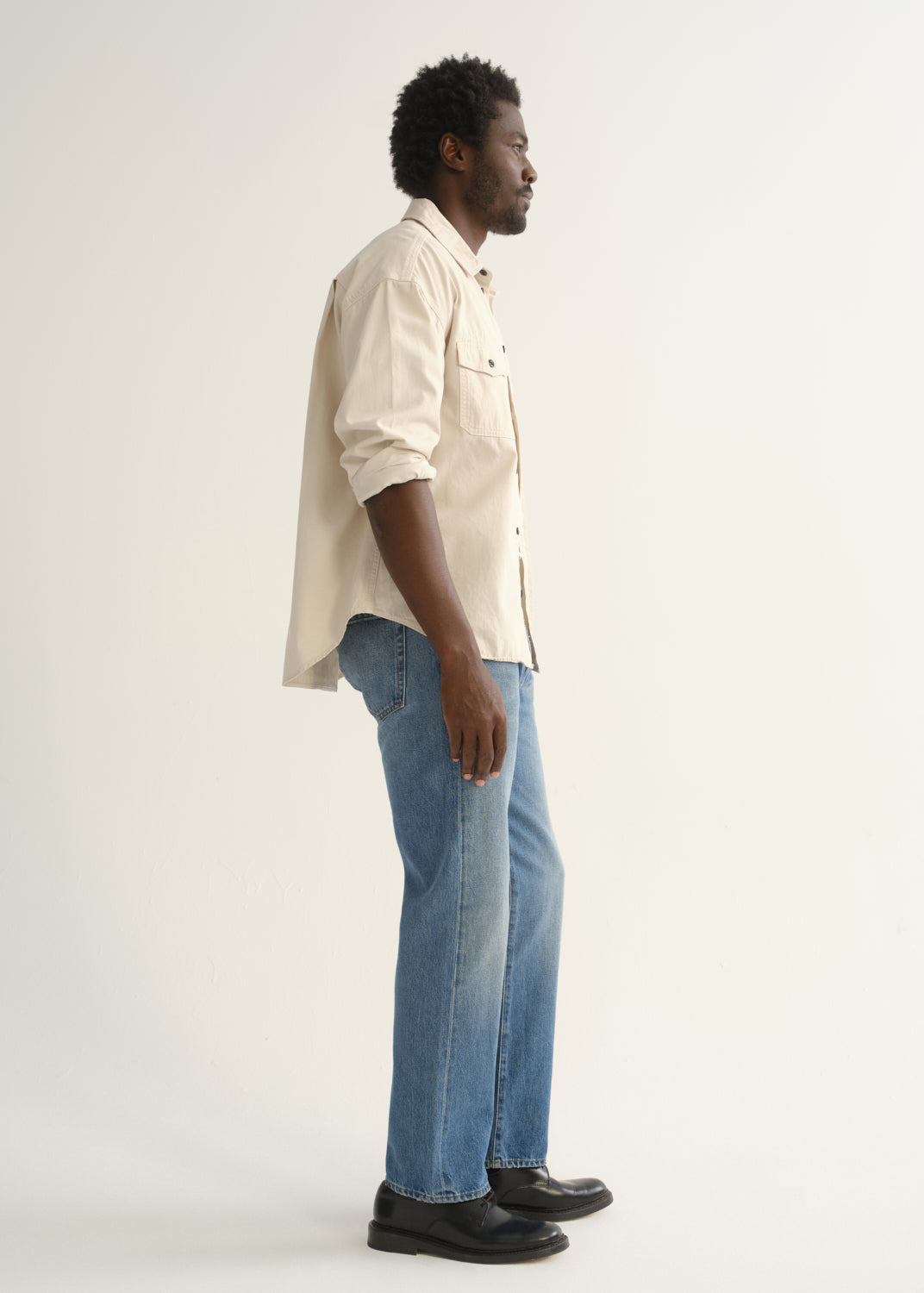 Person wearing light blue straight leg jeans with a cream button-up shirt and black shoes, side full body view on a light grey background.