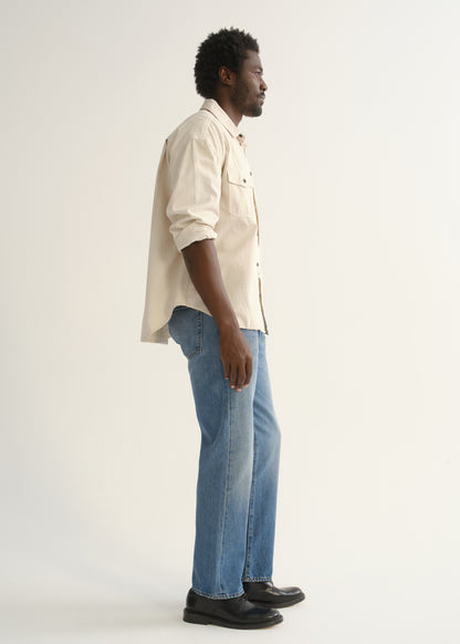 Person wearing light blue straight leg jeans with a cream button-up shirt and black shoes, side full body view on a light grey background.