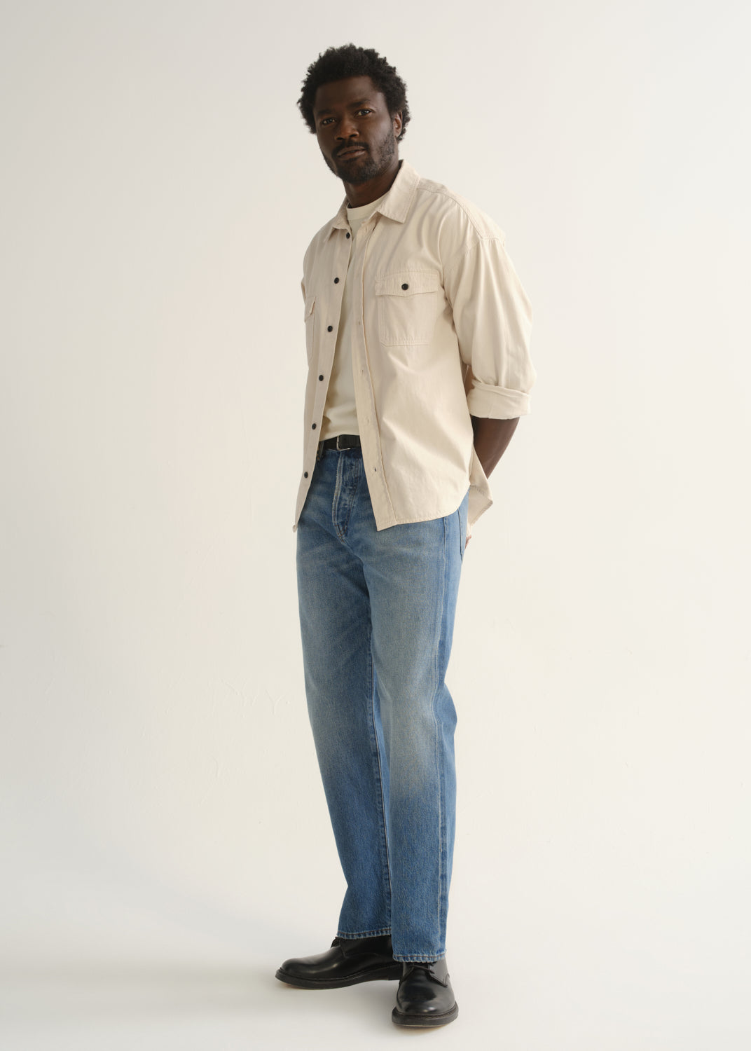 Person wearing light blue straight leg jeans with a cream button-up shirt and black shoes, angled front full body view on a light grey background.