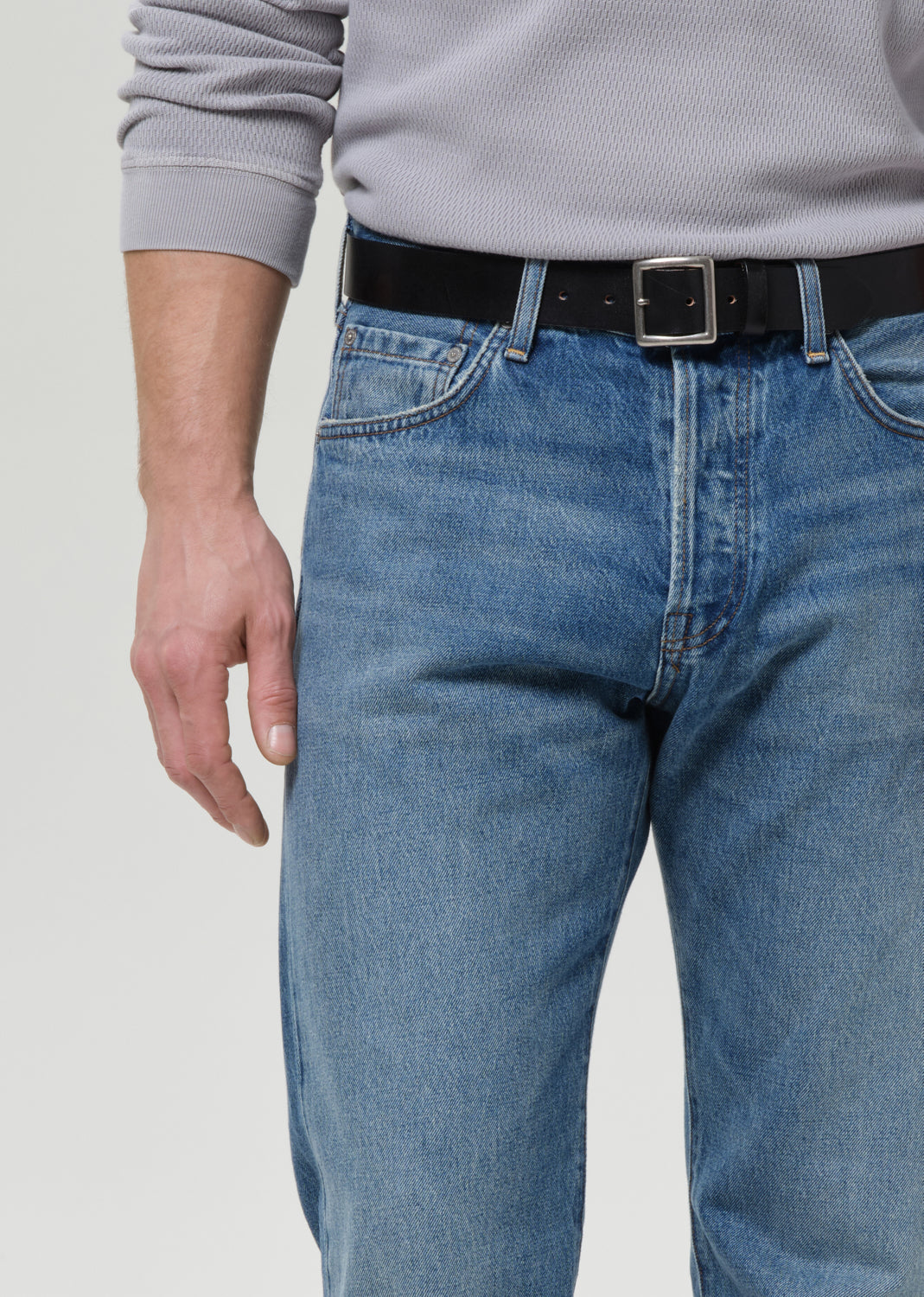 A close-up of a person wearing blue denim jeans with a black belt and a gray ribbed long-sleeve top, cropped from the chest down against a grey background.