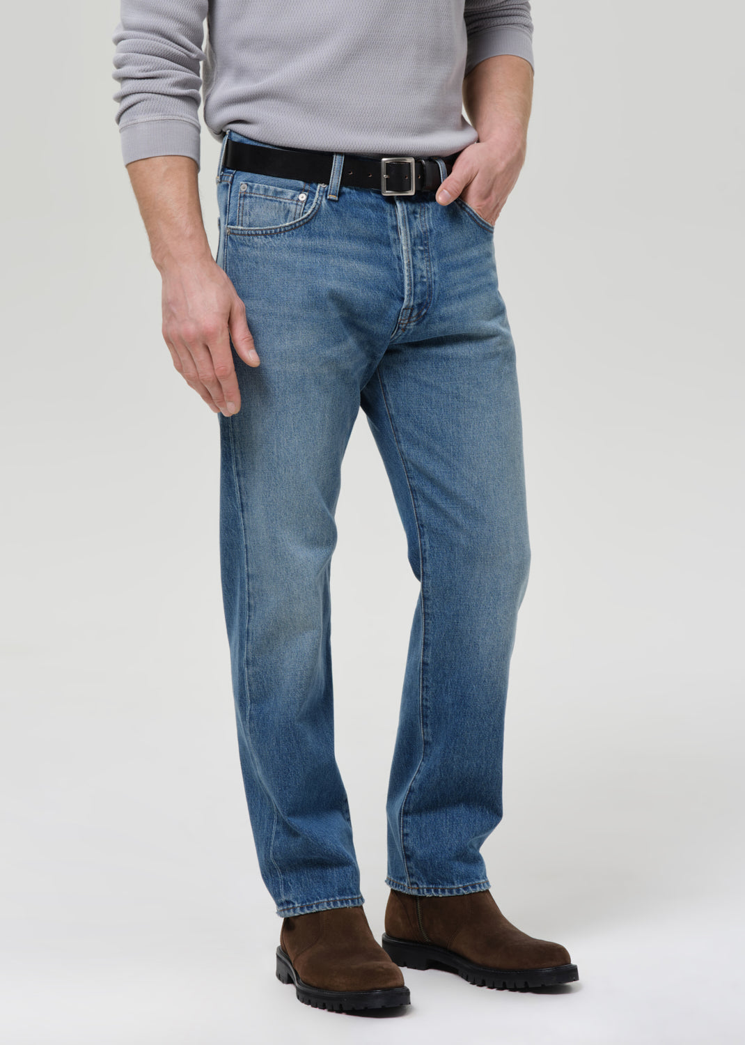A man wearing medium-wash blue jeans with a black belt, a light grey long-sleeve shirt, and brown boots, standing against a grey background.