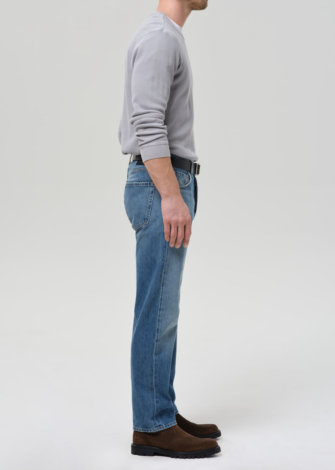 A side profile view of a man wearing blue denim jeans, a light grey long-sleeve shirt, a black belt, and brown suede boots, standing against a grey background.