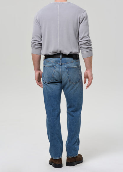 A man viewed from behind wearing a light grey long-sleeve shirt, blue jeans, a black belt, and brown shoes, standing against a grey background.