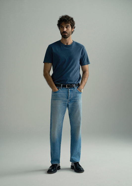 A man wearing a dark blue crewneck t-shirt, light blue jeans, a black belt, and black leather loafers, standing against a neutral gray studio background.