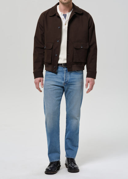 Person wearing a brown jacket, white shirt, blue jeans, and black shoes on a grey background.