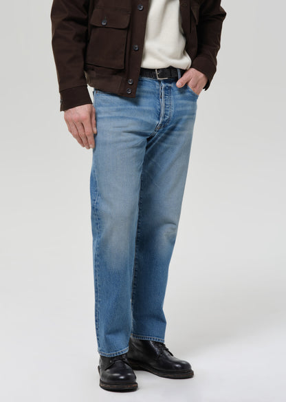 Person wearing a brown jacket, white shirt, blue jeans, and black shoes on a grey background.