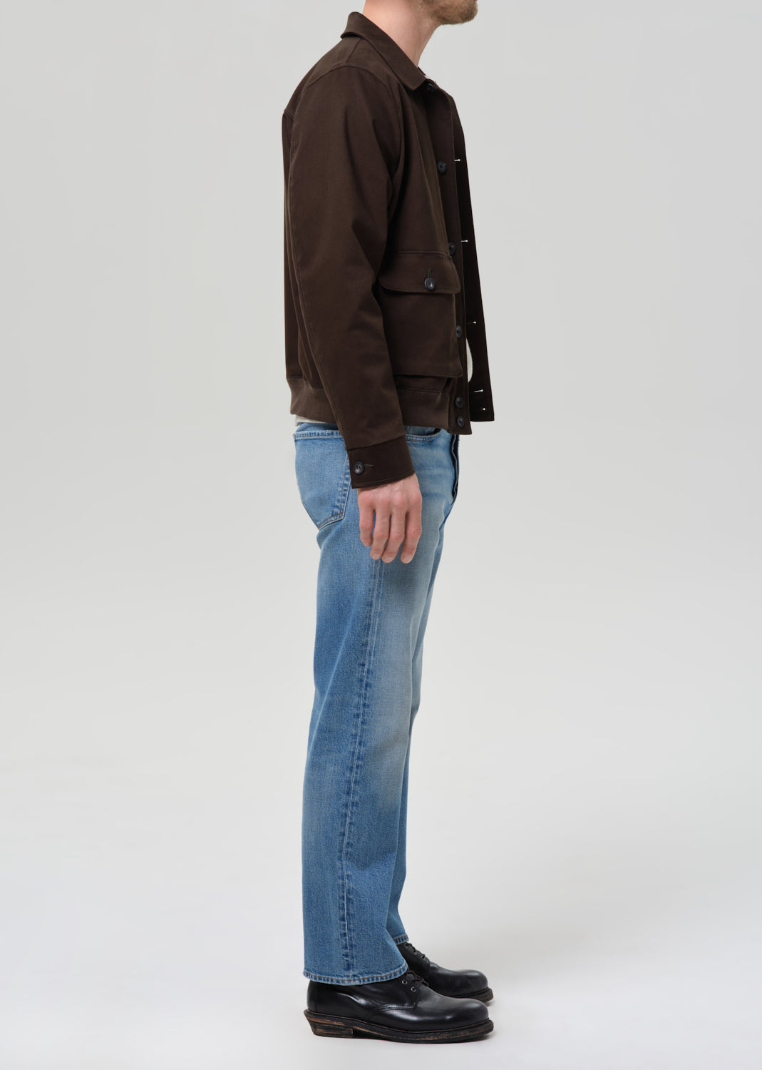 Person wearing a brown jacket and blue jeans on a grey background.