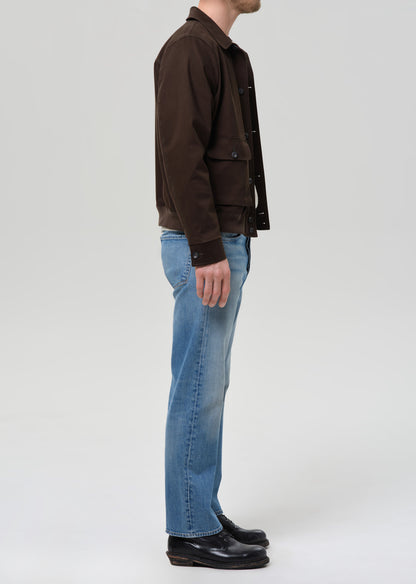 Person wearing a brown jacket and blue jeans on a grey background.