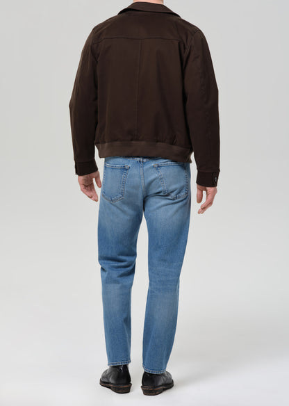 Person wearing a brown jacket and blue jeans on a grey background.