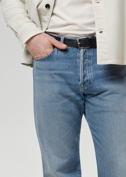 Man wearing a cream shirt, cream tee, blue jeans, and brown shoes on a plain background