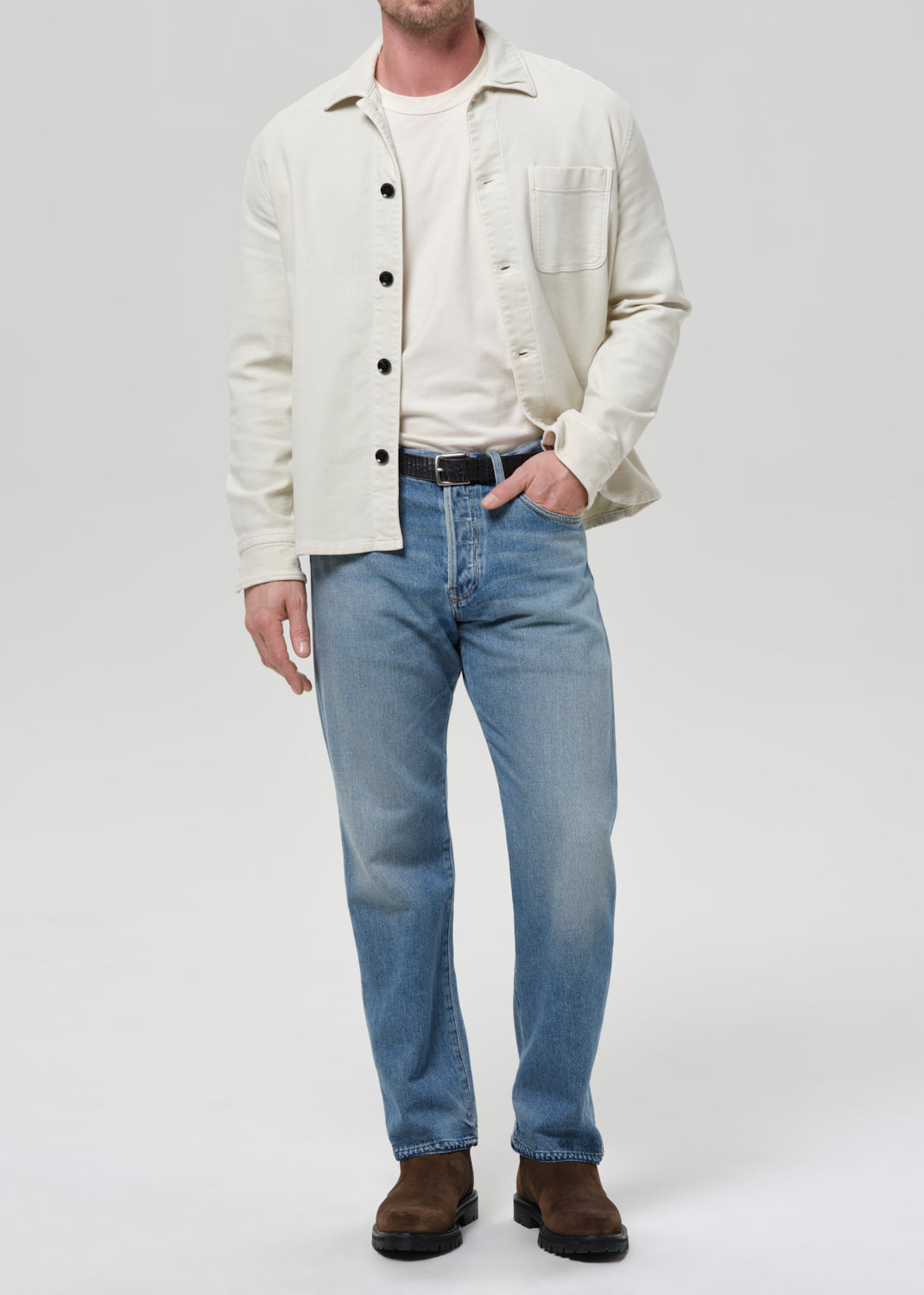 Man wearing a cream shirt, cream tee, blue jeans, and brown shoes on a plain background