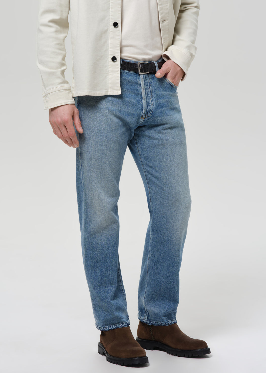 Man wearing a cream shirt, cream tee, blue jeans, and brown shoes on a plain background