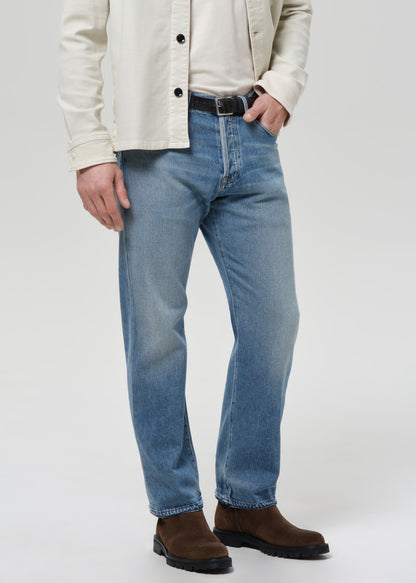 Man wearing a cream shirt, cream tee, blue jeans, and brown shoes on a plain background