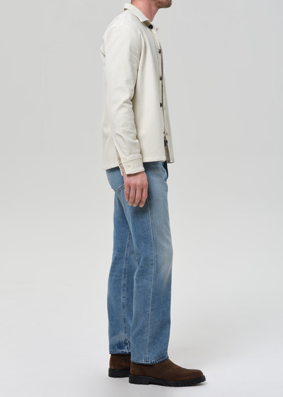 Man wearing a cream shirt, cream tee, blue jeans, and brown shoes on a plain background