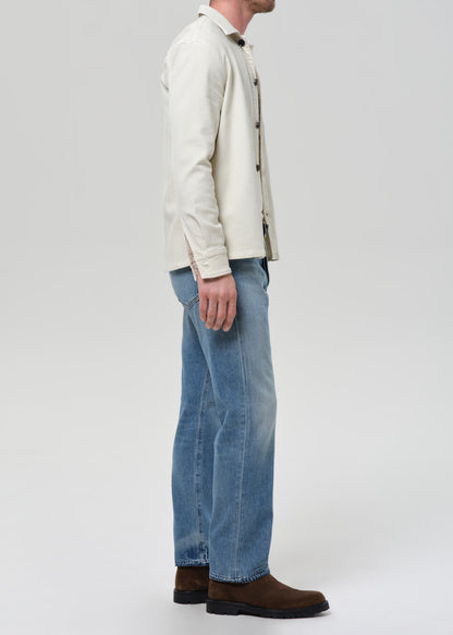 Man wearing a cream shirt, cream tee, blue jeans, and brown shoes on a plain background