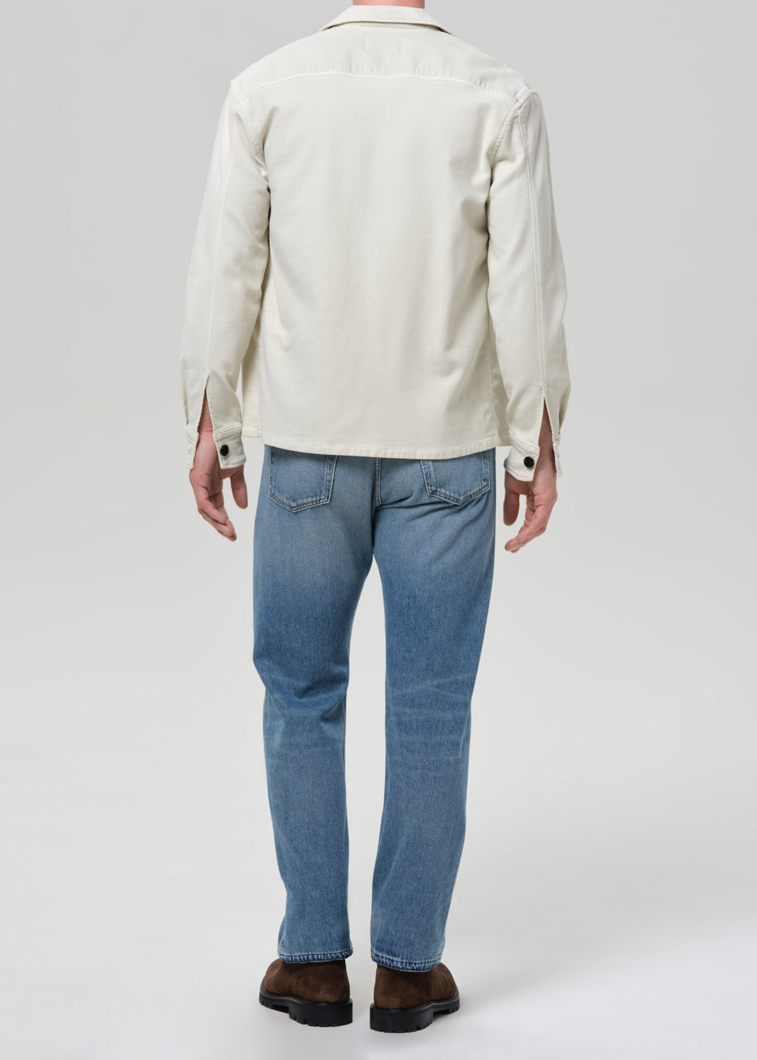 Man wearing a cream shirt, cream tee, blue jeans, and brown shoes on a plain background