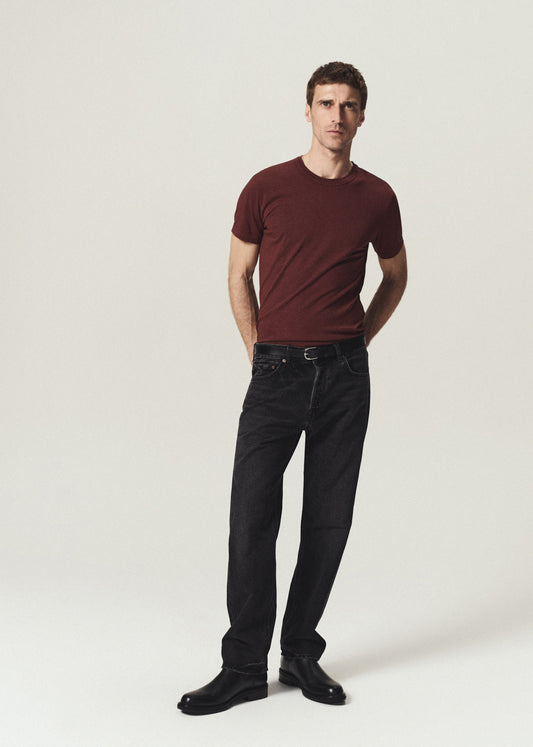 A man modeling a dark maroon crewneck t-shirt, black jeans, and black leather shoes against a plain light gray studio background.