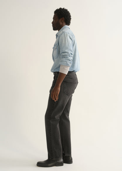 Person wearing washed black straight leg jeans with a light blue denim button-up shirt and black shoes, side full body view on a light grey background.