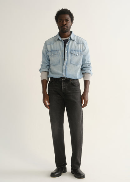 Person wearing washed black straight leg jeans with a light blue denim button-up shirt and black shoes, front full body view on a light grey background.