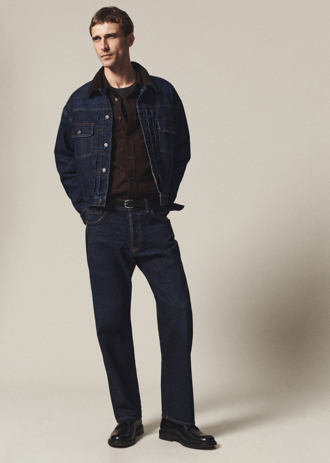 A man modeling a dark blue denim jacket and matching dark blue jeans, styled with a brown shirt, black belt, and black leather shoes against a plain beige studio background.
