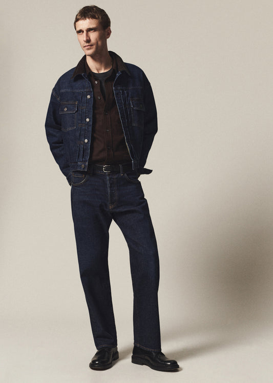 A man modeling a dark blue denim jacket and matching dark blue jeans, styled with a brown shirt, black belt, and black leather shoes against a plain beige studio background.