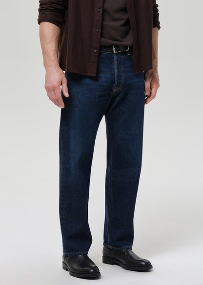 Person wearing dark blue jeans and a brown jacket on a grey background