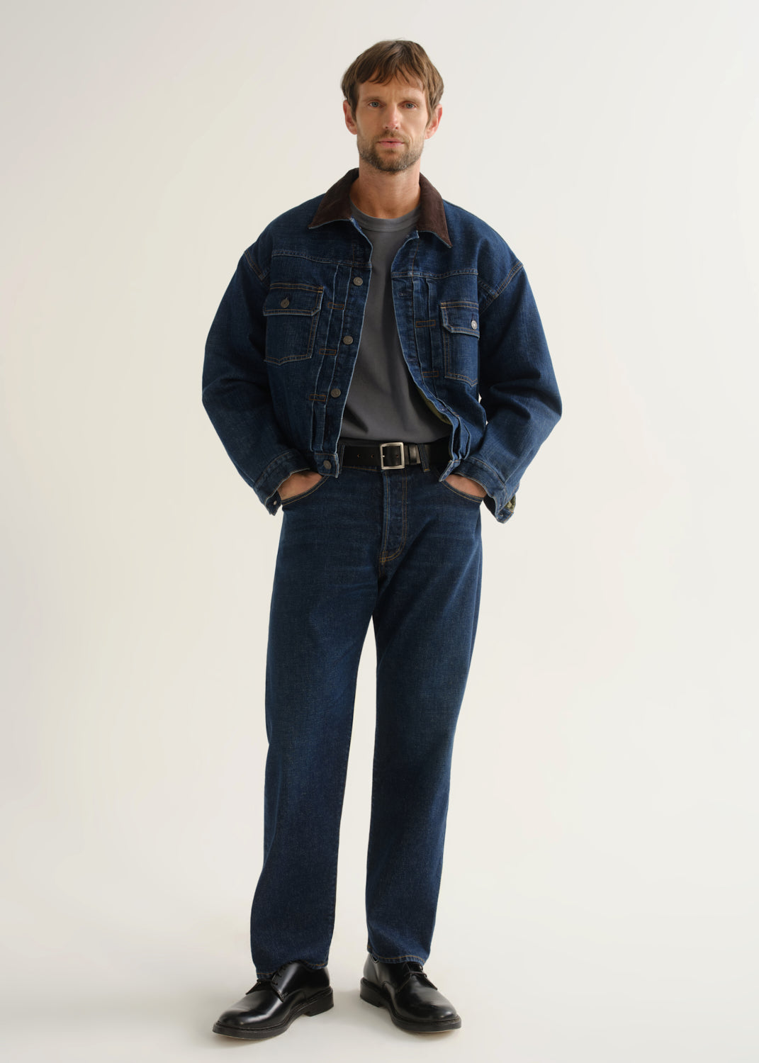 Person wearing dark indigo straight leg jeans with a matching denim jacket and black boots, front full body view on a light grey background.