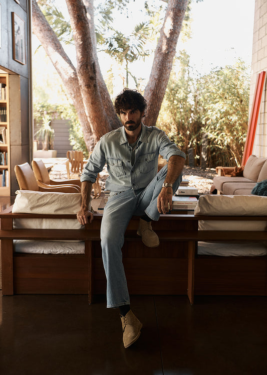 Man sitting on a wooden table in a sunlit outdoor living area with trees and furniture wearing a light wash denim shirt and jeans