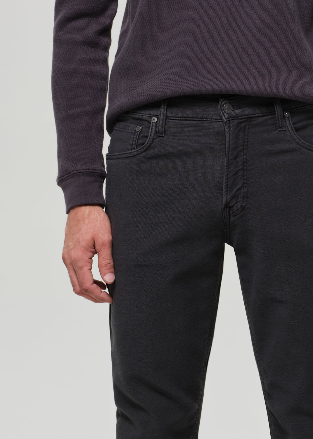 Person wearing a dark crewneck and dark grey pants on a plain background