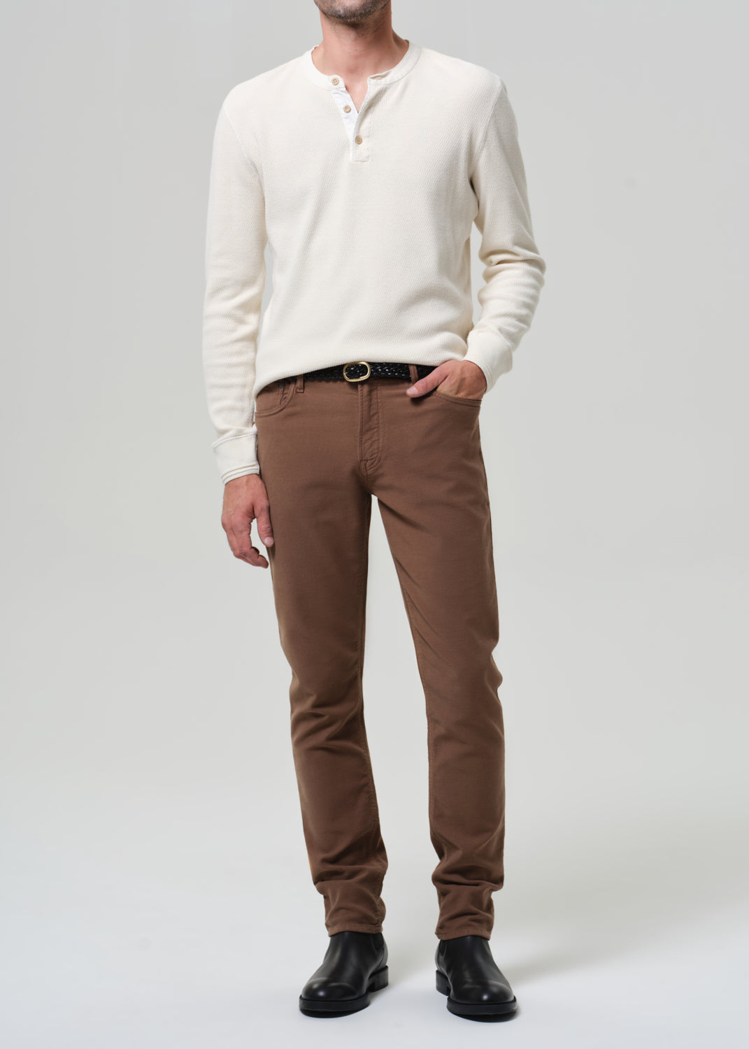 Man wearing a white henley and brown pants on a plain background