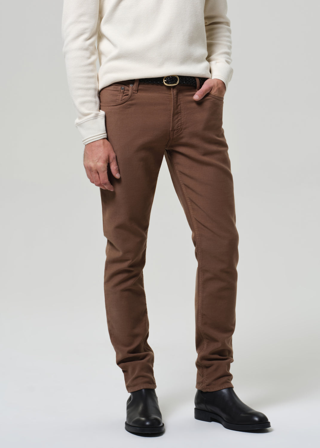 Man wearing a white henley and brown pants on a plain background