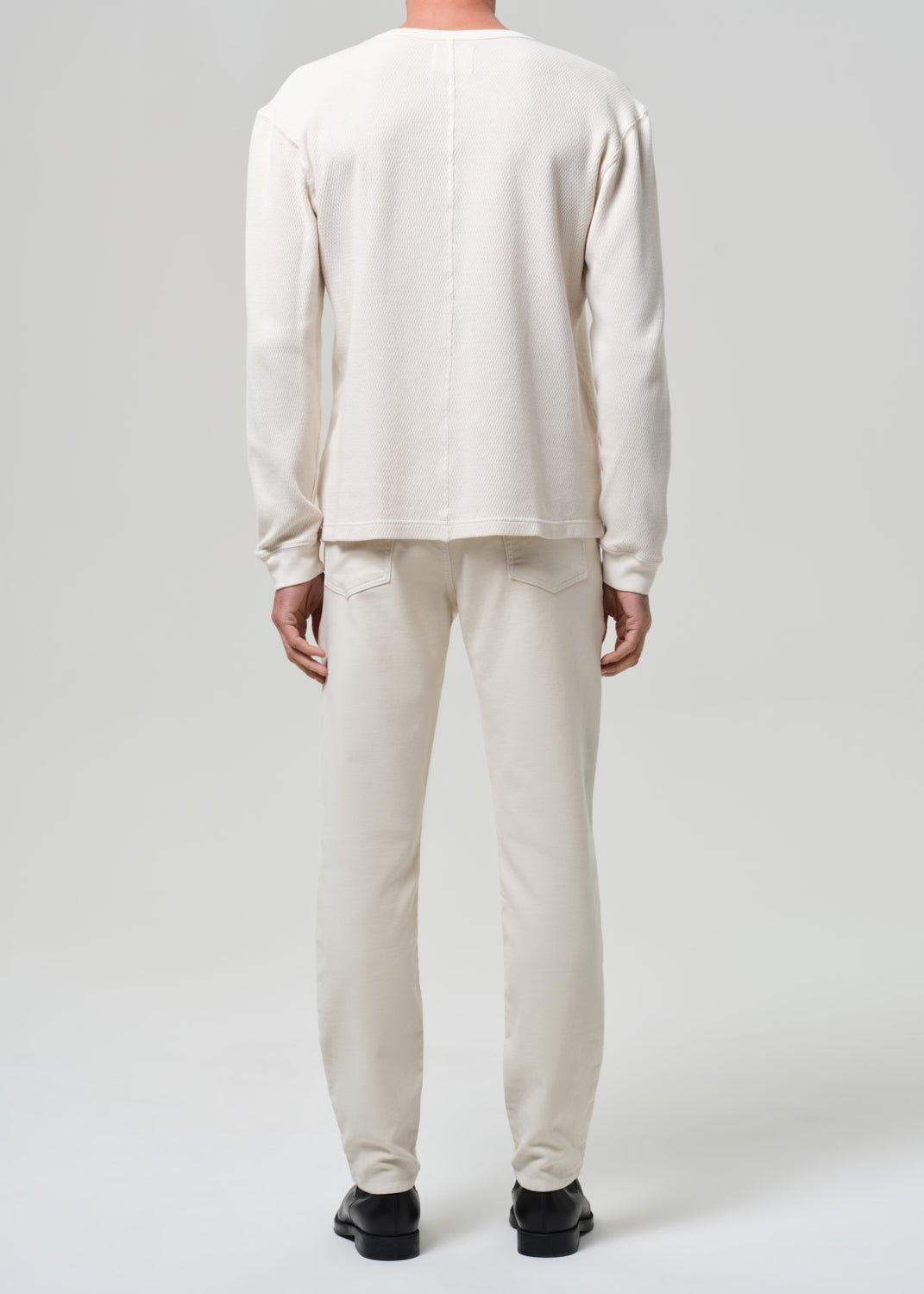 Person wearing a white crewneck and cream pants with black shoes on a plain background