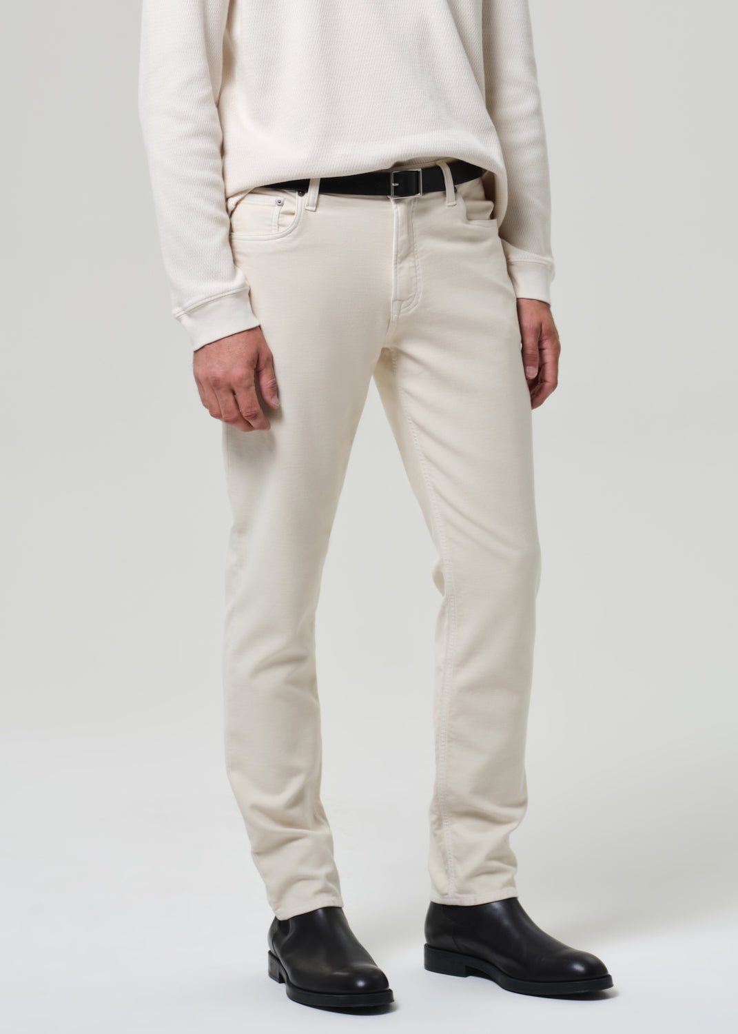 Person wearing a white crewneck and cream pants with black shoes on a plain background