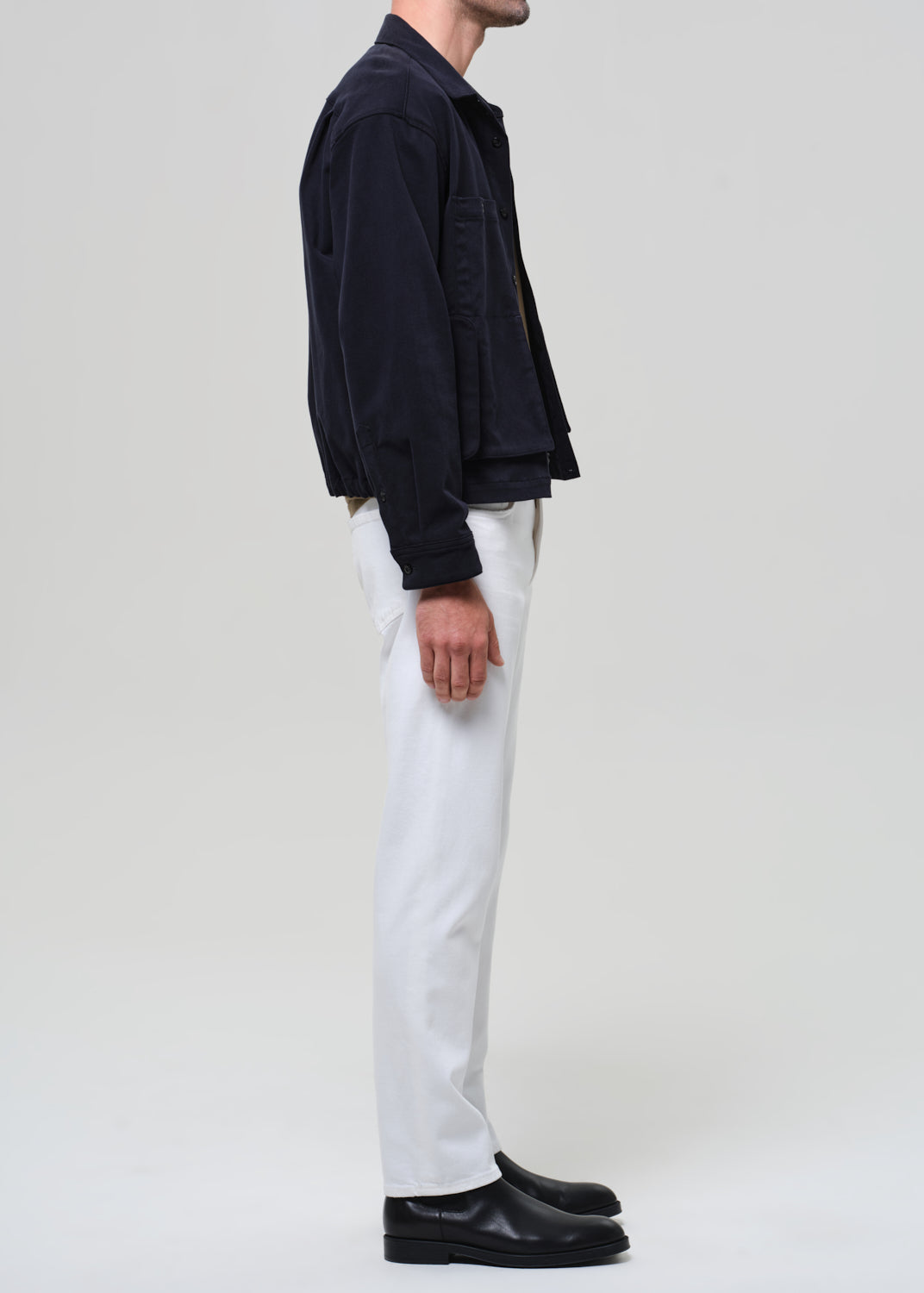 Person wearing a dark jacket, white pants, and black shoes on a plain background