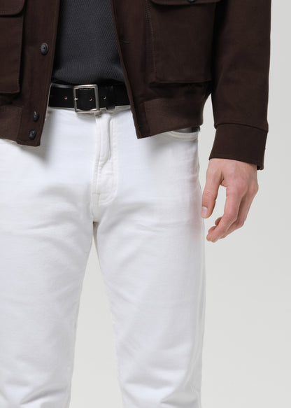 A close-up of a person wearing white pants, a black belt, a dark gray knit top, and a brown jacket, cropped from the chest down against a grey background.