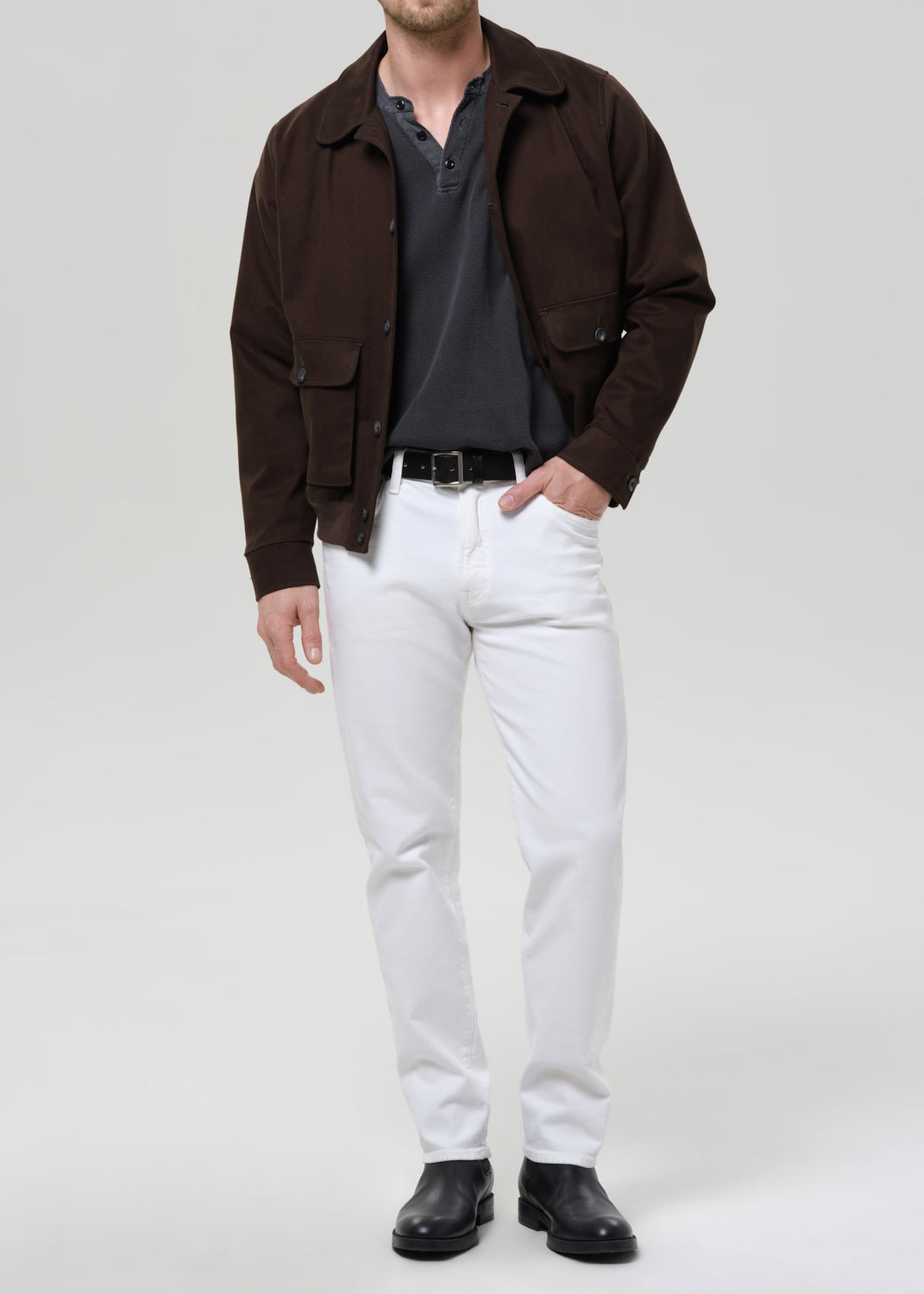 A man modeling a dark brown utility jacket over a dark grey henley shirt, paired with white trousers, a black belt, and black leather boots, against a grey background.