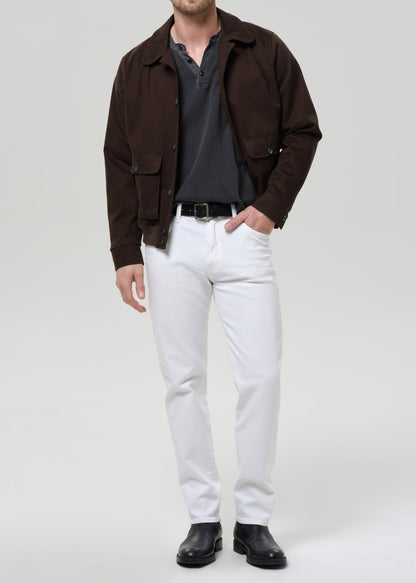 A man modeling a dark brown utility jacket over a dark grey henley shirt, paired with white trousers, a black belt, and black leather boots, against a grey background.