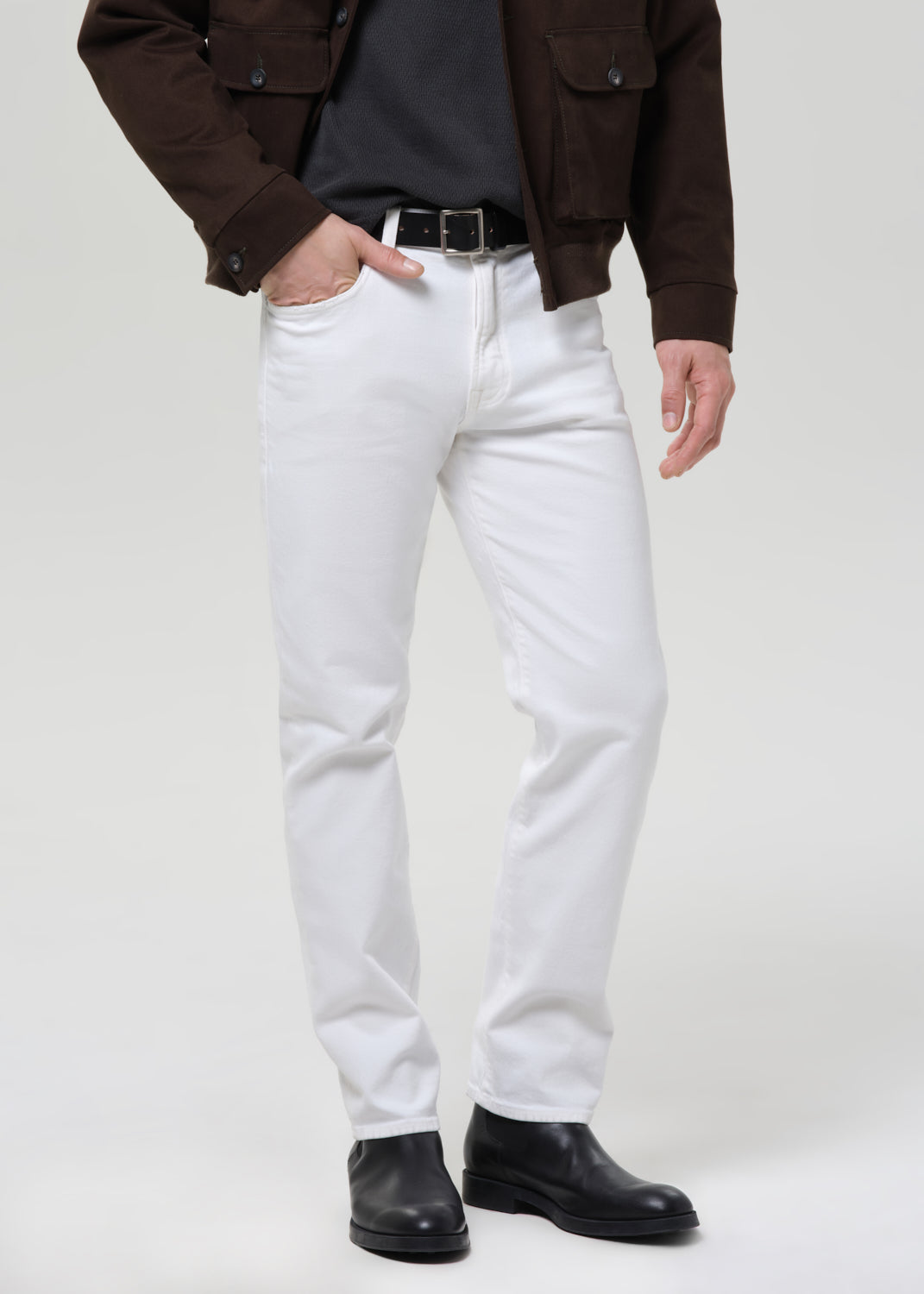 A person wearing white slim-fit jeans with a black belt, paired with a dark brown jacket, black shirt, and black leather boots, standing against a grey background.