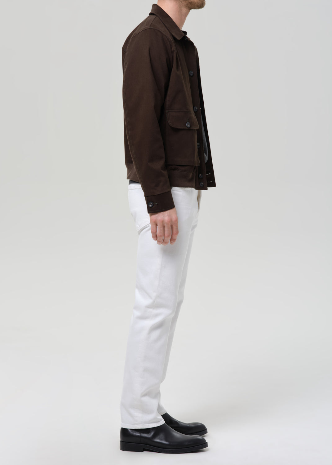 A man shown in profile wearing a dark brown jacket, white trousers, and black shoes against a grey background.
