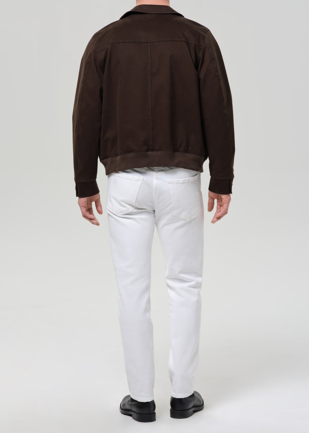 A man viewed from behind wearing a dark brown suede jacket, white trousers, and black shoes, standing against a grey background.