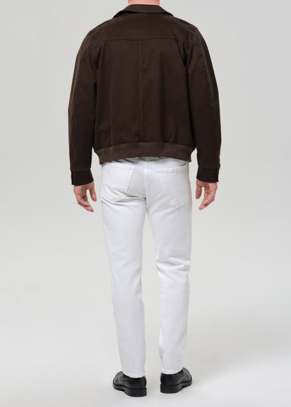 A man viewed from behind wearing a dark brown suede jacket, white trousers, and black shoes, standing against a grey background.