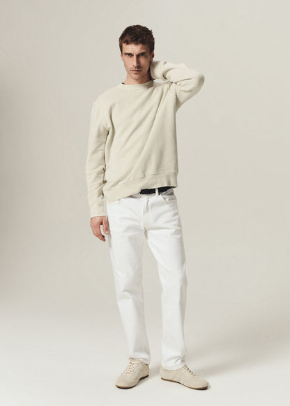 A male model wearing a cream-colored crewneck sweatshirt, white trousers, and matching cream sneakers, standing against a light beige studio background.