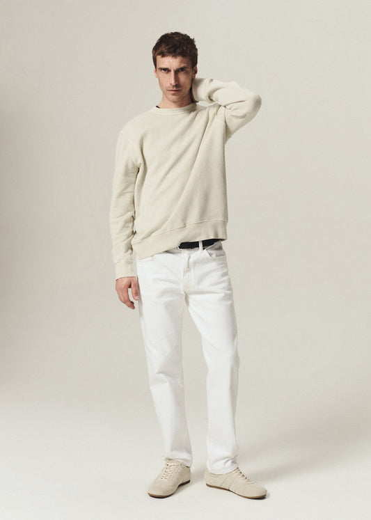 A male model wearing a cream-colored crewneck sweatshirt, white trousers, and matching cream sneakers, standing against a light beige studio background.
