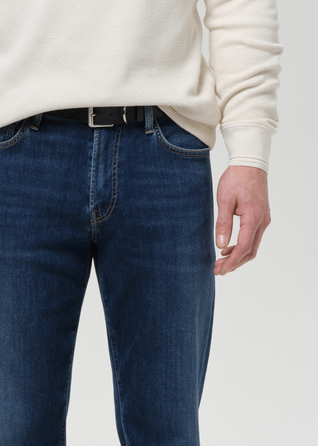 A close-up of a person wearing dark blue denim jeans with a black belt and a cream ribbed knit sweater, cropped from the chest down against a grey background.