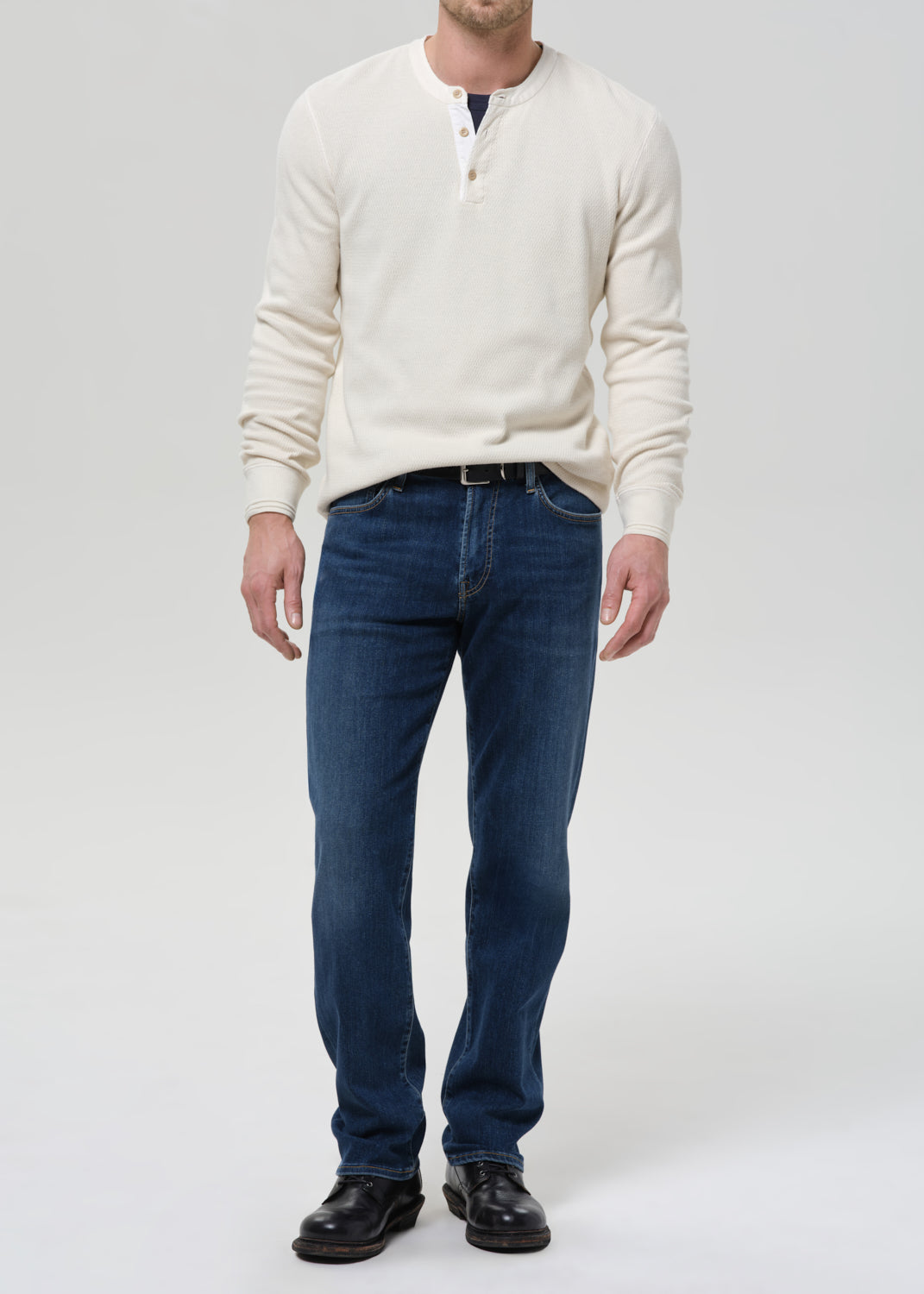 A man wearing a cream-colored long-sleeve henley sweater, dark blue jeans, and black leather shoes, standing against a grey background.
