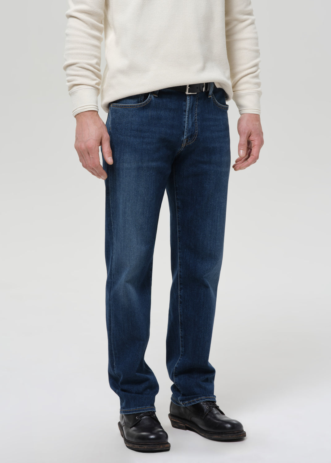 A person wearing dark blue straight-leg jeans, a cream sweater, and black leather shoes, standing against a grey background.