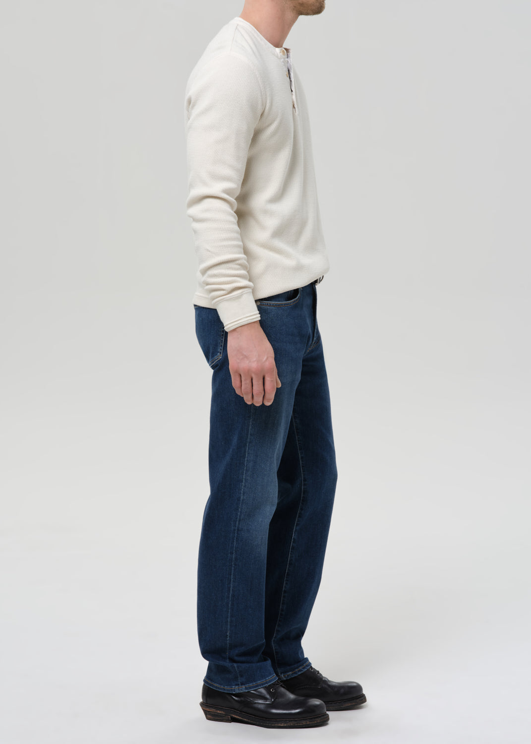 A man shown in profile wearing dark blue jeans, a cream long-sleeve henley shirt, and black leather shoes against a grey background.
