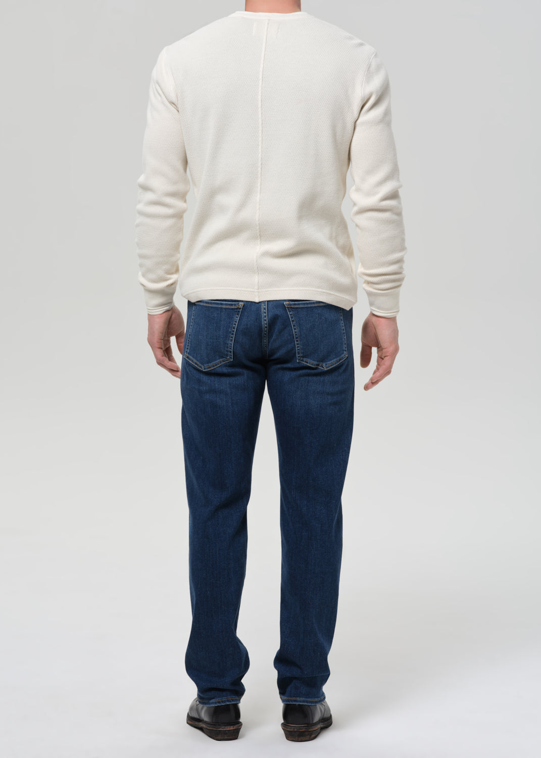 A man viewed from the back wearing a cream-colored long-sleeve knit sweater and dark blue jeans, standing against a grey background.