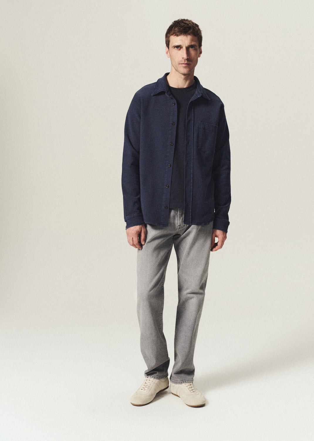 A man modeling a dark navy blue overshirt over a black t-shirt, paired with light gray jeans and off-white sneakers, standing against a plain light gray studio background.